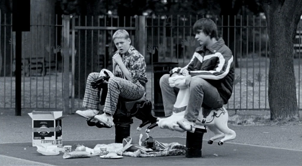 Somers Town (2008) movie still: fullbody — A public playground area features a metal fence and trees in the background. Two teenage boys sit on…