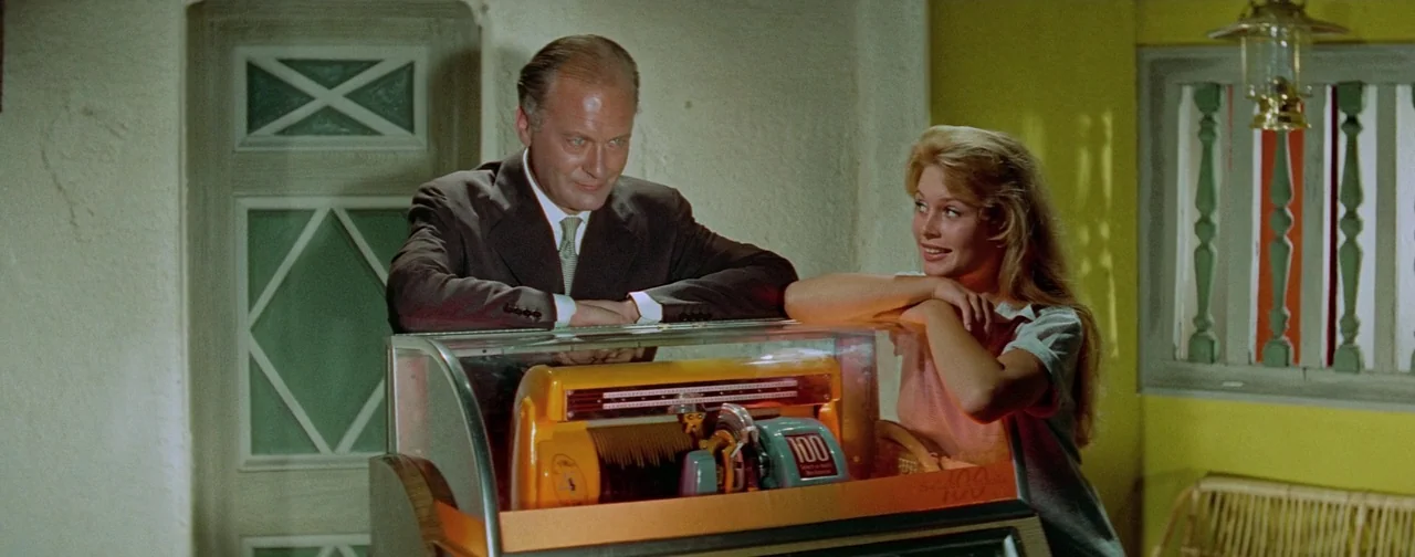 And God Created Woman (1956) movie still: medium — An interior space features bright yellow walls and a wooden door with diamond panels. A man and a…