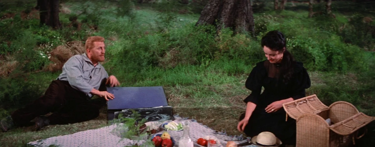 Lust For Life (1956) movie still: wide — A grassy meadow surrounded by dense trees hosts a picnic blanket with assorted food items. A man sits by an…