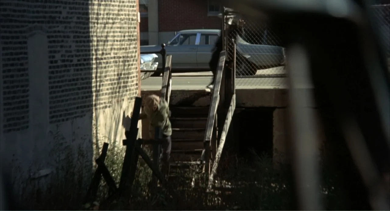 Cooley High (1975) movie still: wide — A neglected loading dock area sits beside a weathered brick wall and overgrown weeds. A man in light clothing…