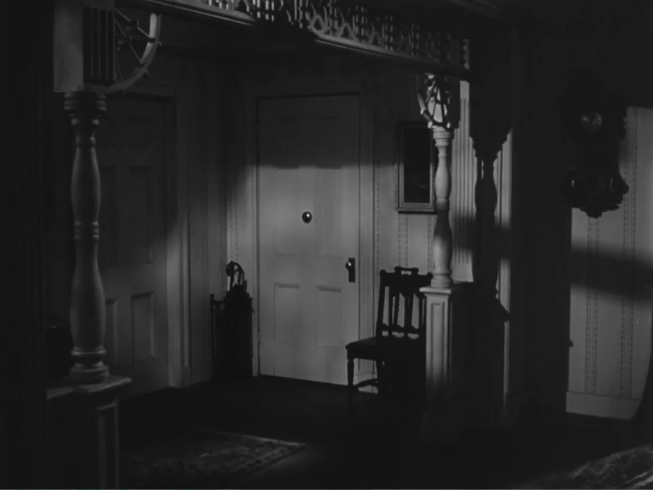 House by the River (1950) movie still: medium — A residential hallway with wooden architectural trim and a door in the center. An empty chair sits…