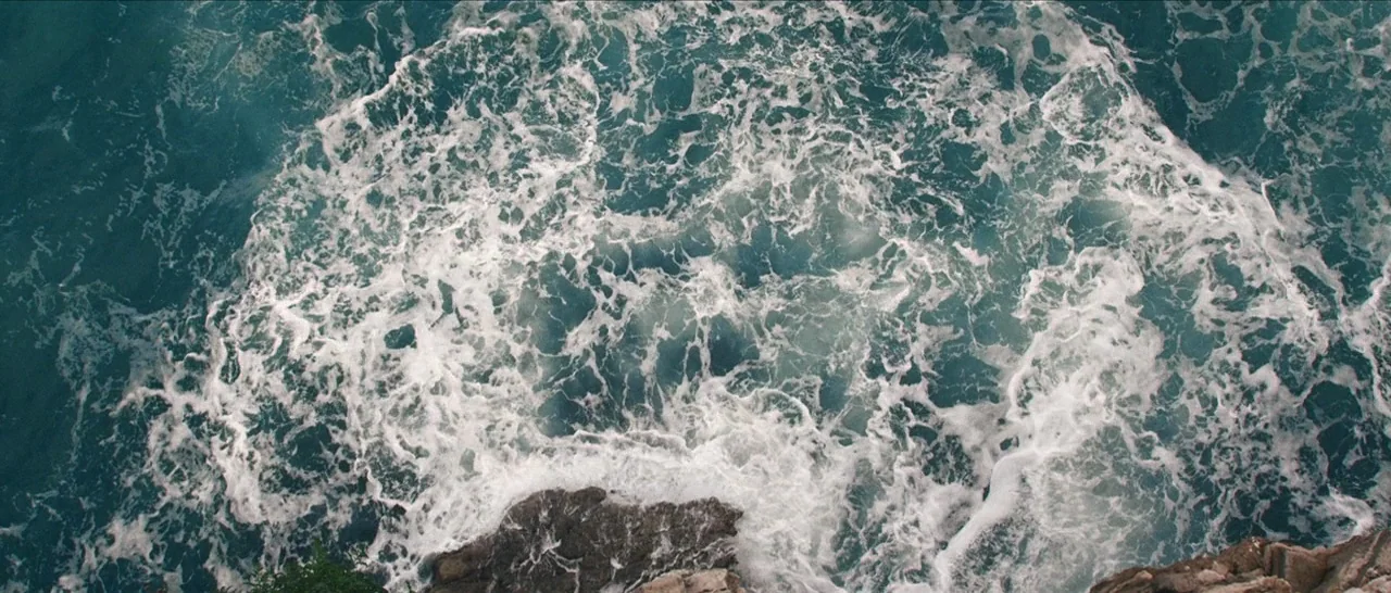 Spring (2014) movie still: establishing — A top-down view shows dark ocean water crashing against the edges of craggy rocks. White sea foam churns…
