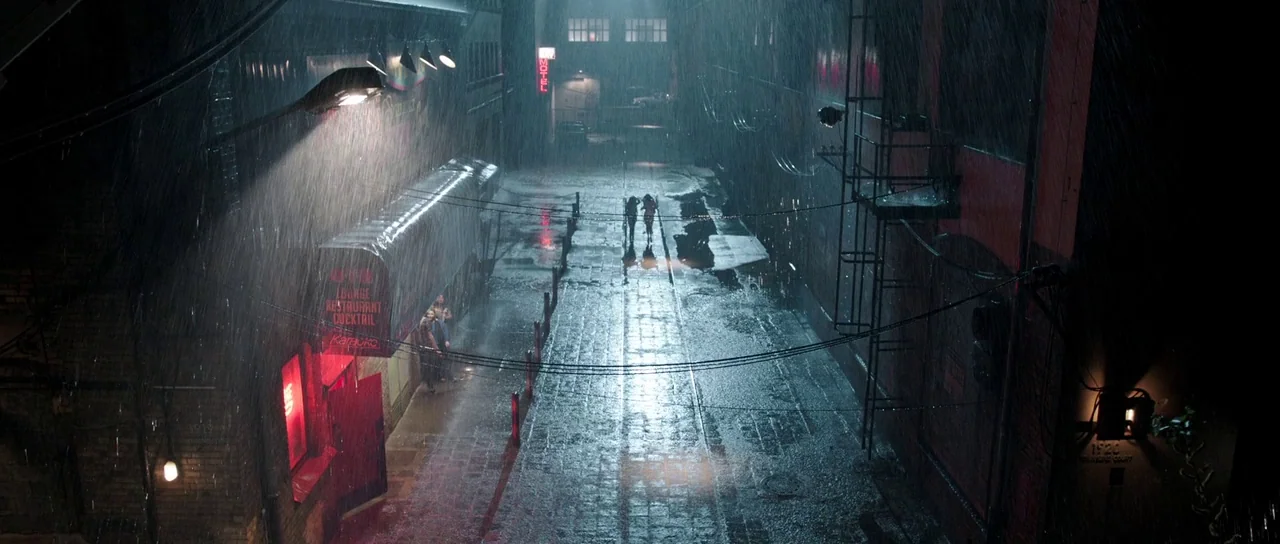 The Voices (2014) movie still: establishing — A dark, wet city street is illuminated by harsh streetlamps and glowing red neon. Rain pours down over…