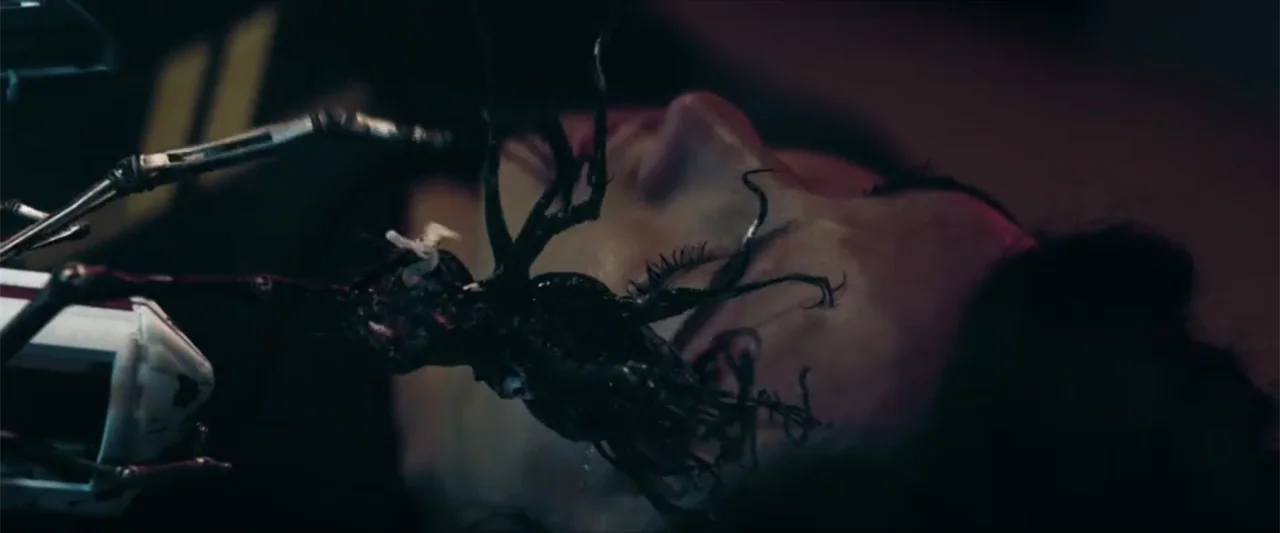 Ash (2025) movie still: closeup — A person lies unconscious on a dark surface in a dim, metallic environment. An amorphous, vein-like black mass is…