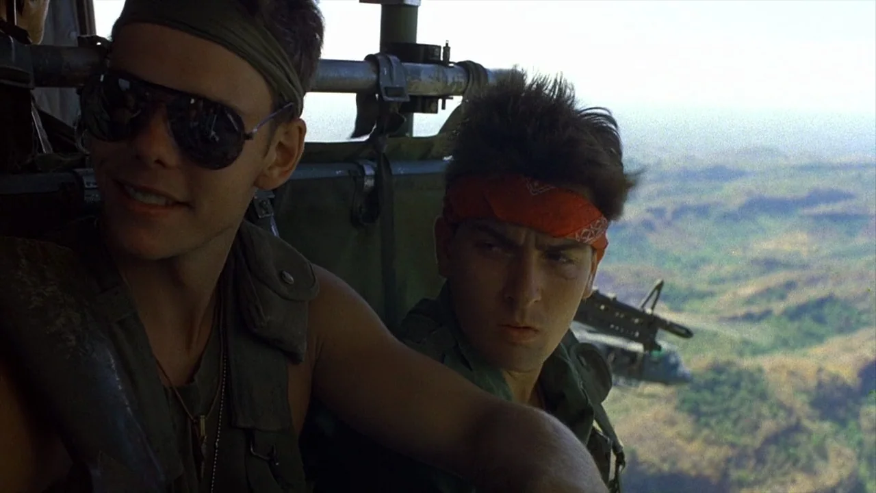 Platoon (1986) movie still: medium — Two men sit inside the open bay of a military helicopter in flight. A vast arid landscape extends far below the…