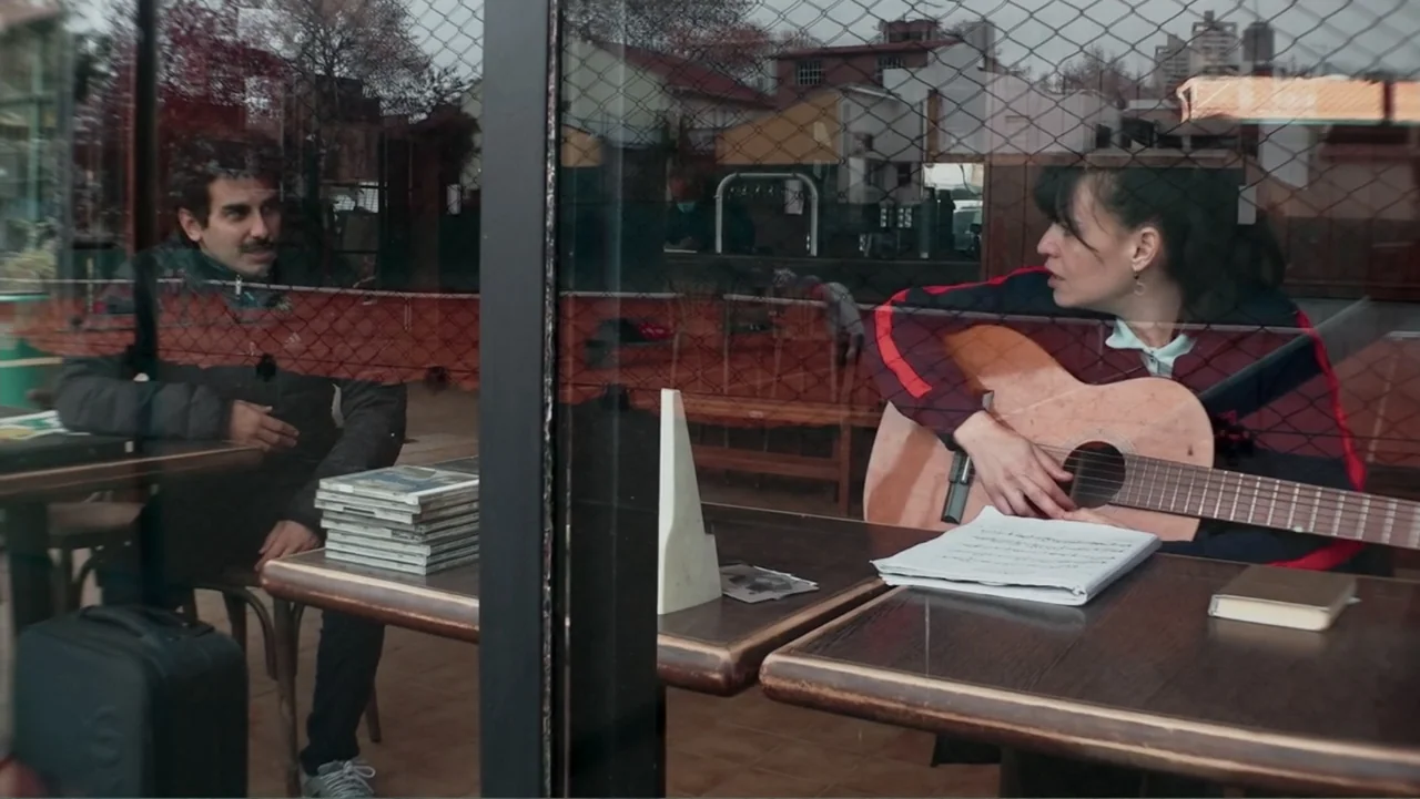 Smog in Your Heart (2022) movie still: medium — A cafe window displays a man sitting at a table opposite a woman playing guitar. Reflections from…