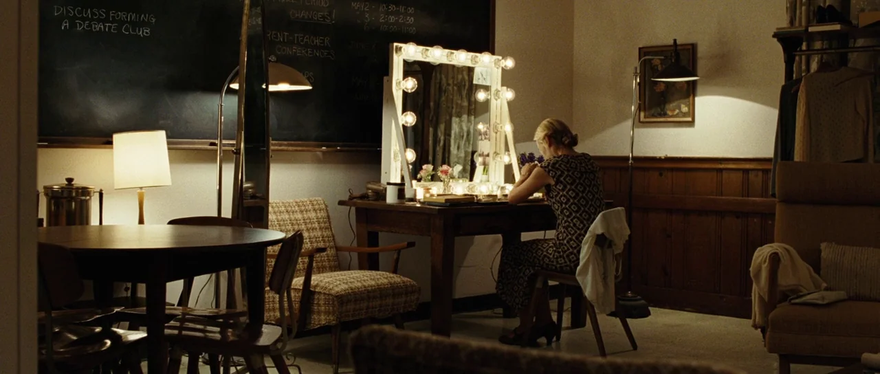 Revolutionary Road (2008) movie still: wide — A dimly lit room functions as a dressing area with a large vanity mirror. A woman sits on a chair,…
