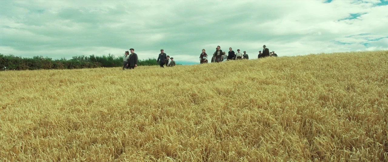 Sunset Song (2015) movie still: wide — A vast golden wheat field stretches under a wide, cloudy sky at midday. A group of men, women, and children…