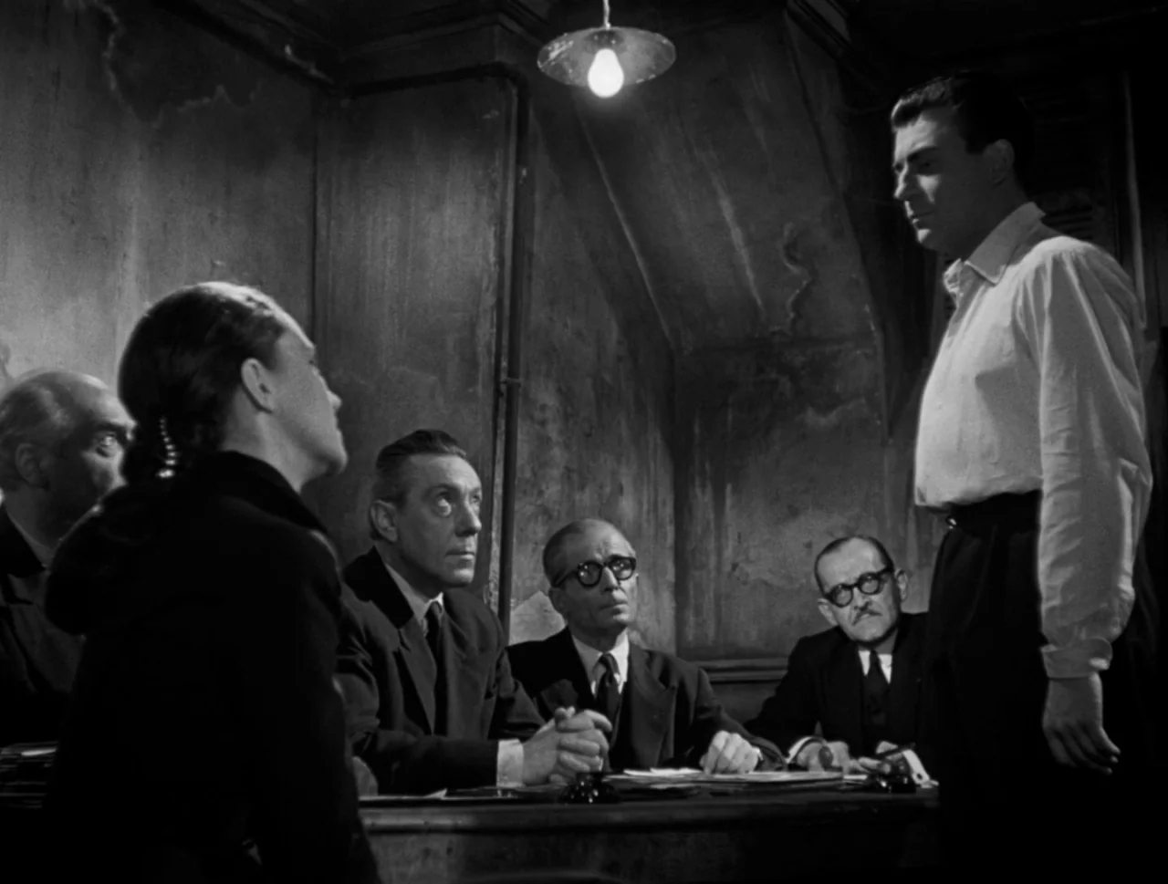 Orpheus (1950) movie still: medium — A stark, dimly lit room features a worn table occupied by four seated men. A lone standing man addresses the…