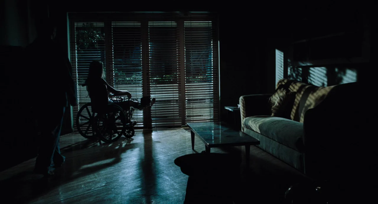 Amores Perros (2000) movie still: wide — A dark living room with hardwood floors is illuminated by moonlight through slatted blinds. A person sits…