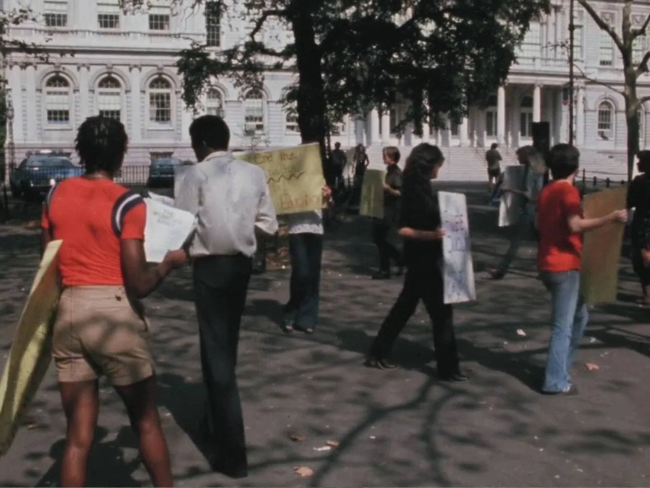 Born in Flames (1983) movie still: fullbody — A public plaza sits in front of a large neoclassical building with columns. A group of people walks…