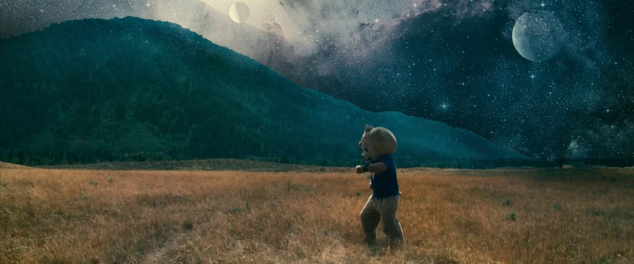 Brigsby Bear (2017) movie still: wide — A dry, grassy landscape meets a dramatic night sky filled with stars and planets. A person in a large bear…