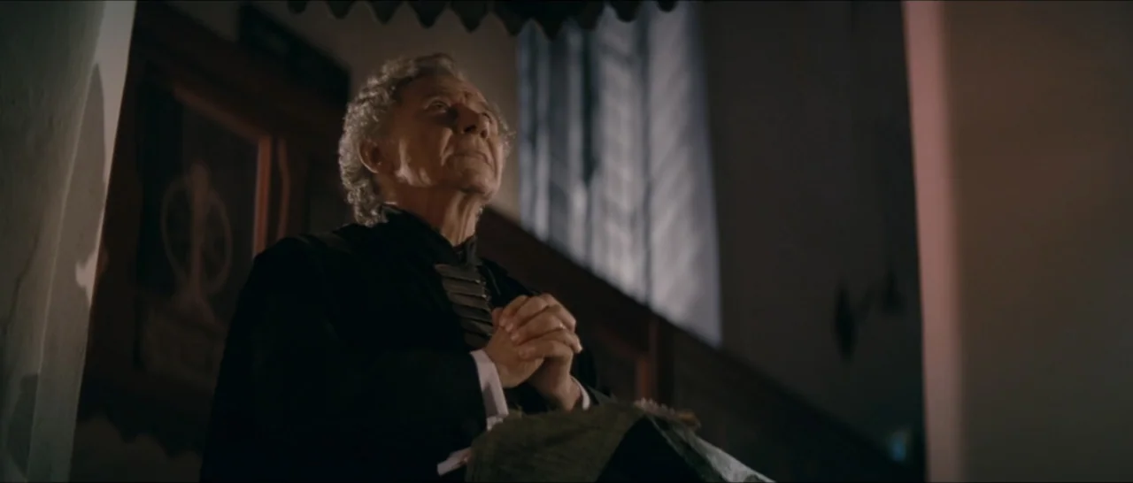 A Farewell to Fools (2013) movie still: medium — A dimly lit religious interior features a stone wall and a window. An elderly man in a formal black…