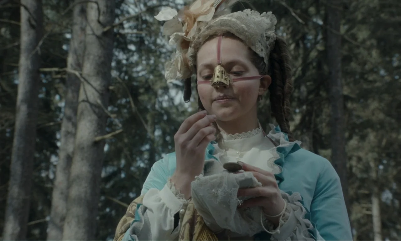 The Ugly Stepsister (2025) movie still: medium — A woman in historical attire stands within a dense forest of evergreen trees. She carefully holds a…