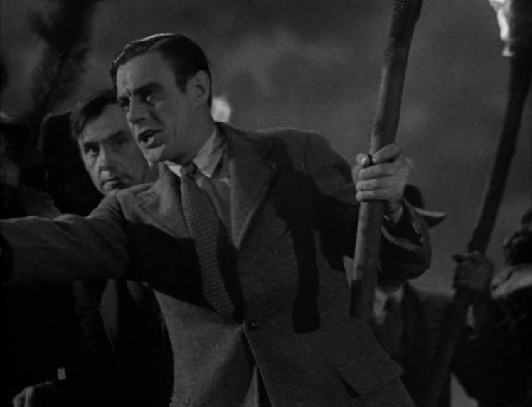 Frankenstein movie still: medium — A dark, nocturnal scene in an open field filled with a crowd of people. A man in a blazer leads the group while…