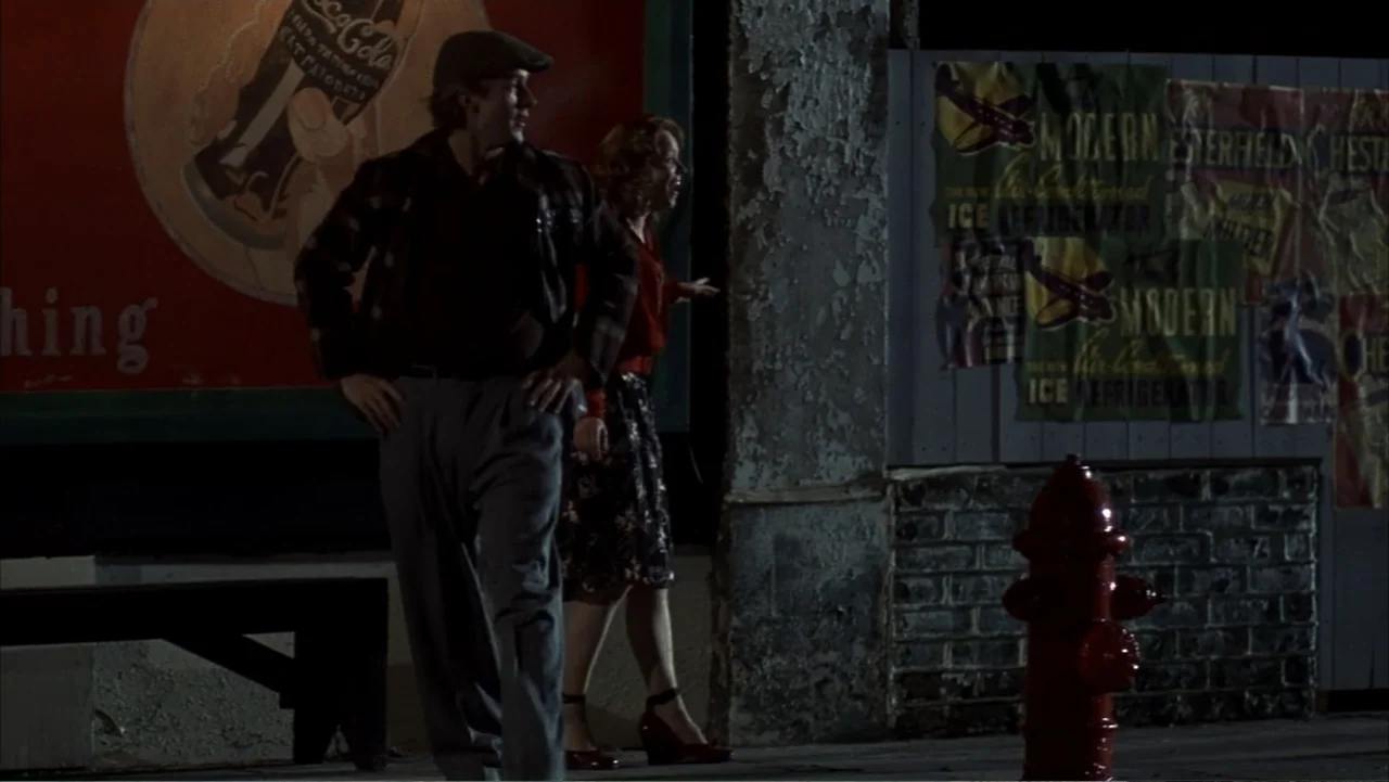 The Notebook (2004) movie still: fullbody — A nighttime city sidewalk features a weathered brick wall adorned with vintage advertising posters. A…
