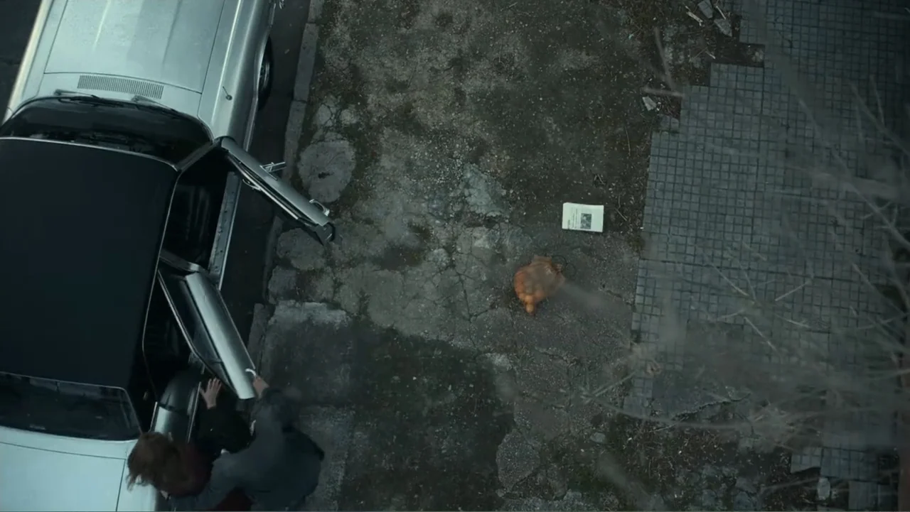 Transfers (2024) movie still: wide — A paved lot at night features a silver car with open doors. A man and a woman stand by the vehicle near a…