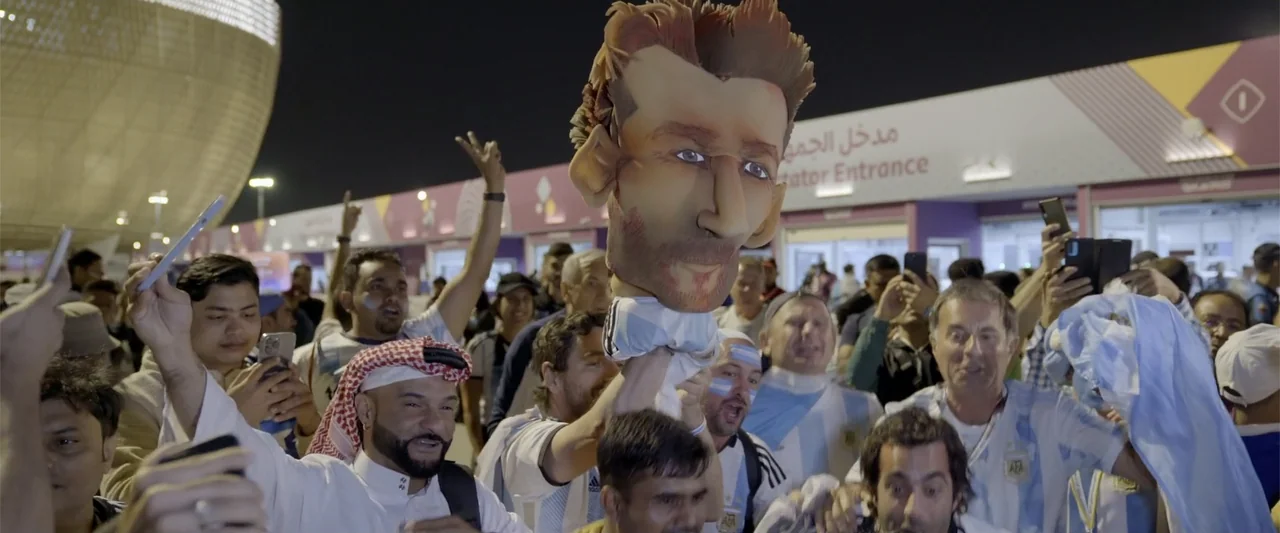 I Choose to Believe (2023) movie still: medium — A dense crowd of cheering soccer fans stands outside a stadium at night. A man holds a large…