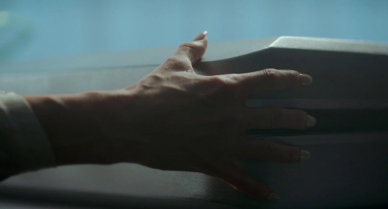 Flux Gourmet (2022) movie still: closeup — A close-up view shows a hand with light-colored fingernails resting on a grey object. The object has a…