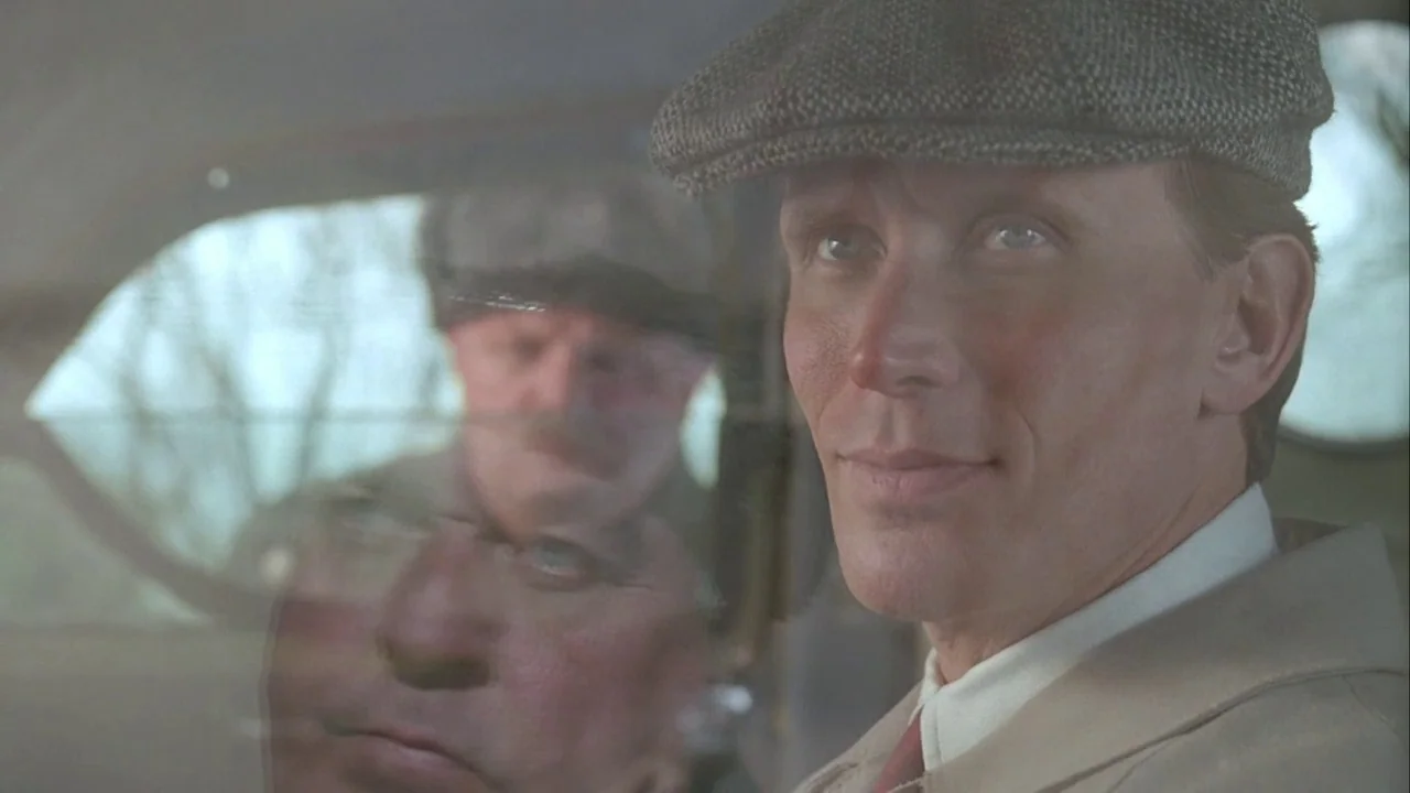 Naked Lunch (1991) movie still: closeup — The interior of a vintage automobile with a view through the window. A man wearing a flat cap looks…