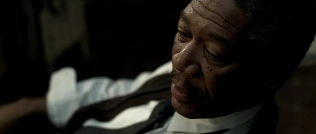 Se7en (1995) movie still: closeup — A dimly lit room with muted earth-toned shadows obscuring the background. An elderly man in a dress shirt and…