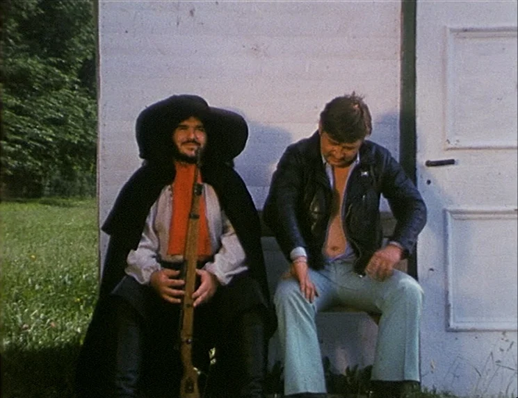 The Niklashausen Journey (1970) movie still: fullbody — Two men sit side-by-side against a white wooden structure at the edge of woods. A man in a…