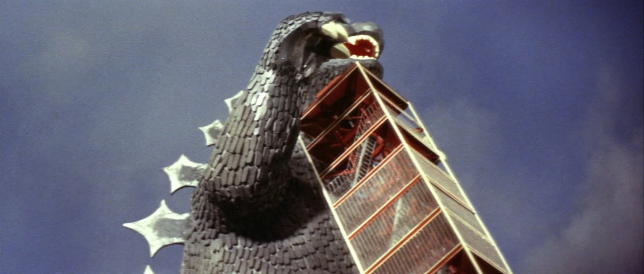 Godzilla Vs Gigan (1972) movie still: medium — A giant reptilian kaiju monster looms against a plain blue daytime sky. The creature grips a tall red…