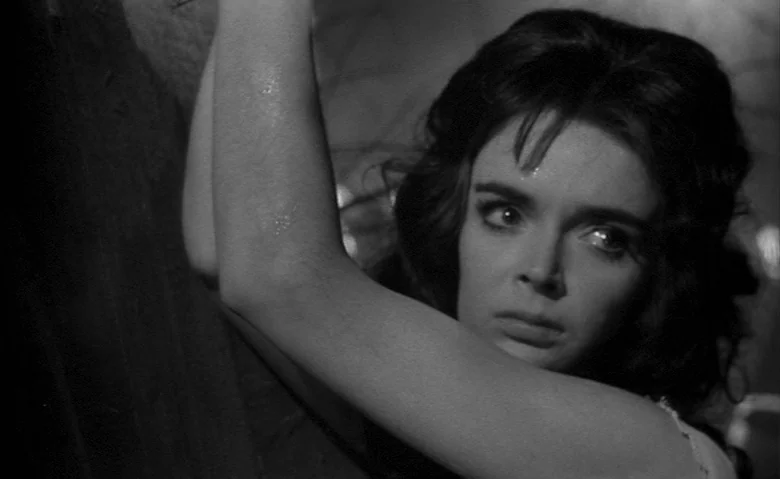 Black Sunday (1960) movie still: closeup — A dark, textured stone wall serves as the backdrop in a dim space. A young woman with dark hair leans…
