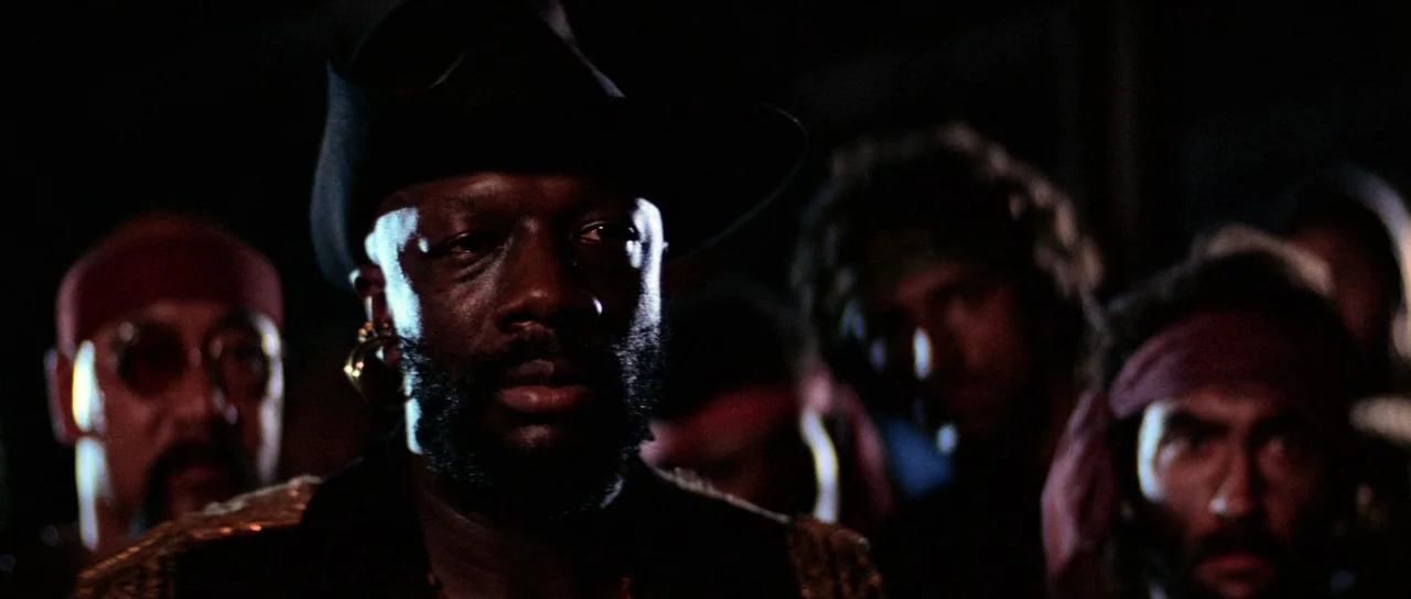 Escape From New York (1981) movie still: medium — A group of men in dimly lit conditions look toward the camera in silence. The central figure wears…