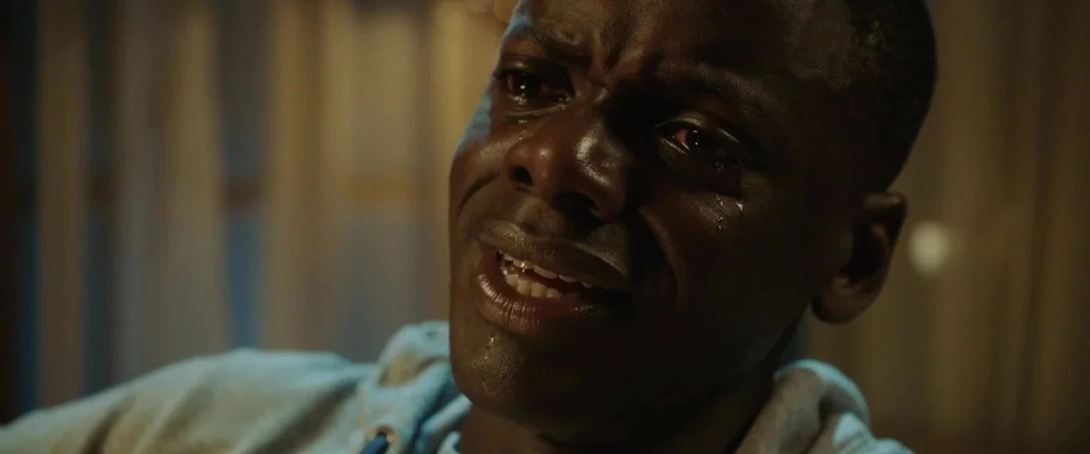 Get Out (2017) movie still: closeup — A dimly lit room provides a dark background for the close-up shot. A young man with tear-streaked skin looks…
