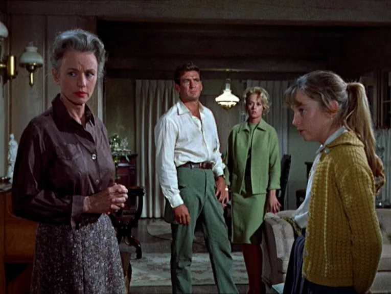 The Birds movie still: medium — A domestic interior with beige curtains, paneled walls, and a warm, lived-in aesthetic. Four people stand in the…