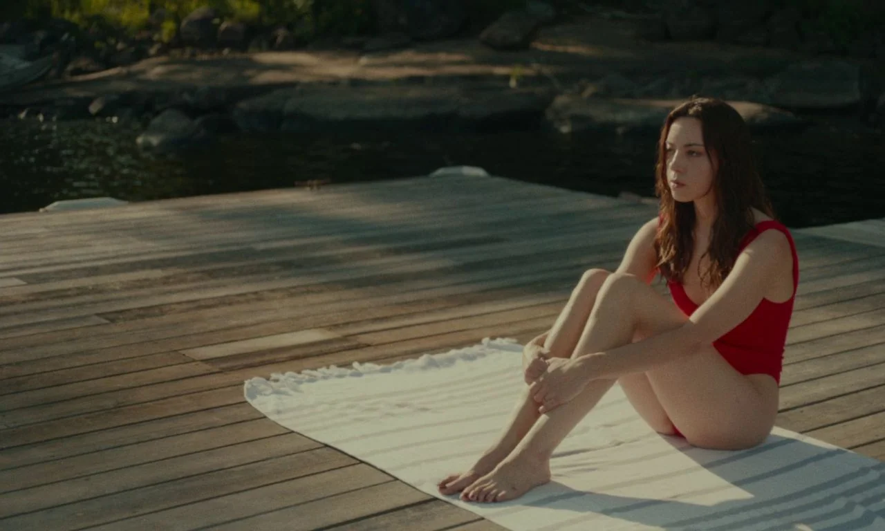 Black Bear (2020) movie still: fullbody — A wooden dock stretches out over dark water towards a rocky shoreline with trees. A woman in a red…