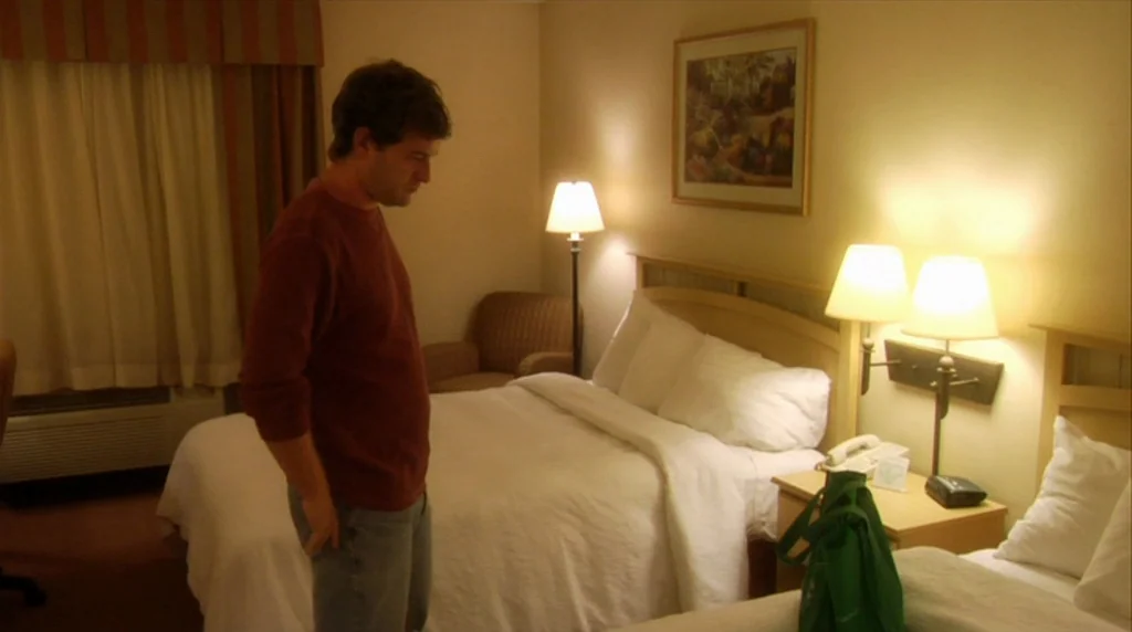 Humpday (2009) movie still: medium — A standard hotel room features white bedding, warm lamps, and striped curtains. A man stands near the bed with…
