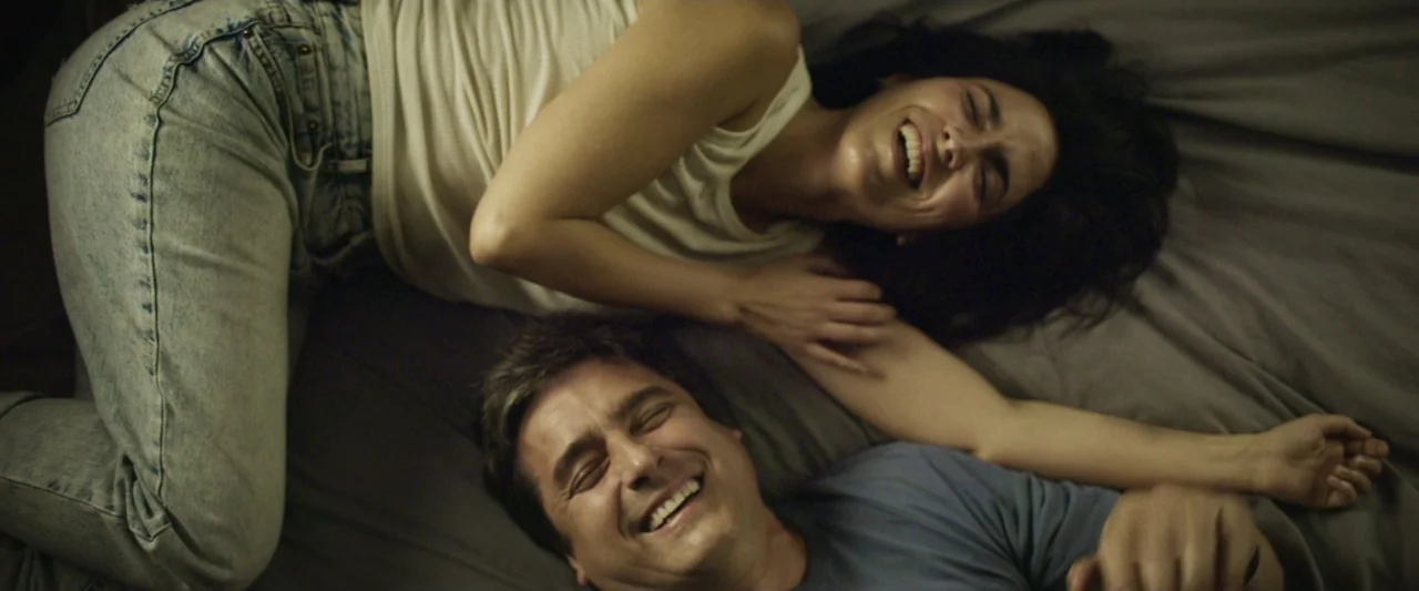 Dog Life (2024) movie still: medium — A man and woman lie together on a grey bed sheet. The two subjects look up at the camera while laughing during…