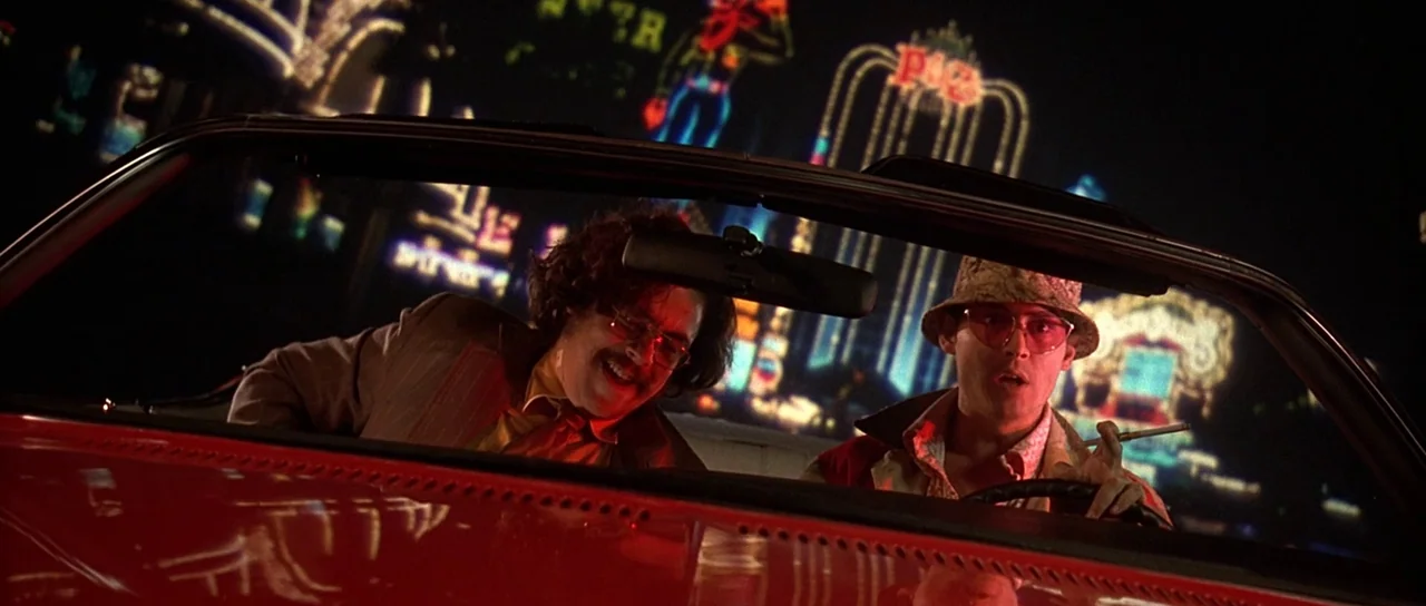 Fear and Loathing In Las Vegas (1998) movie still: medium — Two men are in the front seat of a red convertible driving at night. A bright Las Vegas…