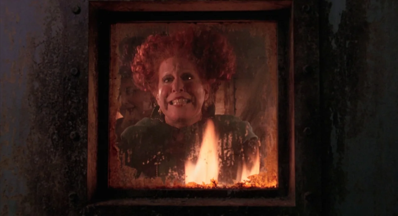 Hocus Pocus (1993) movie still: closeup — A woman stands behind a glass pane set into a rusted metal frame. Intense flames lick the bottom of the…