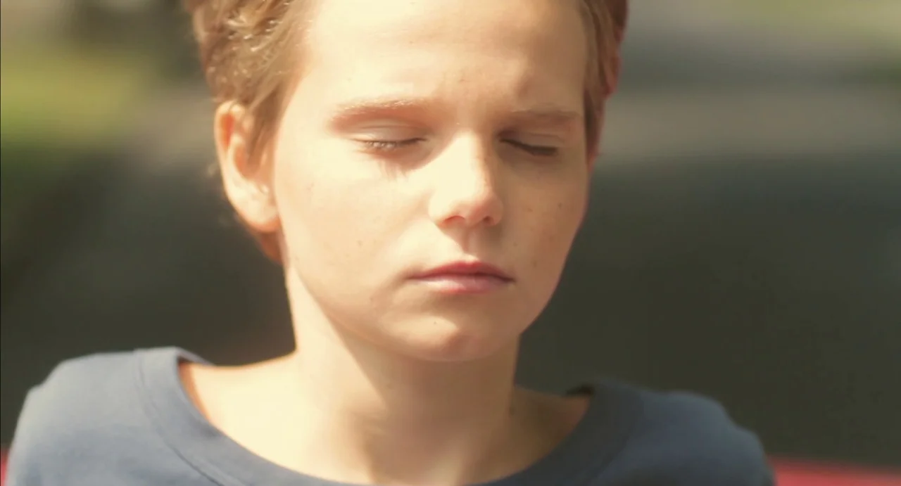 Tomboy (2011) movie still: closeup — A close-up shot of a young person outside in daylight. The subject keeps their eyes closed with a calm facial…