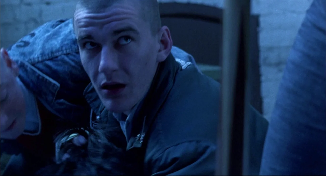 Romper Stomper (1992) movie still: closeup — A dimly lit basement features brick walls and cold blue-toned artificial light. Two young men with…