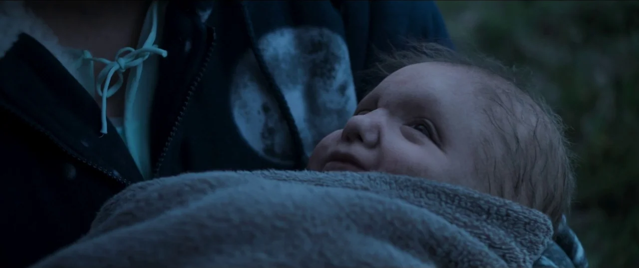 Border movie still: closeup — A dark outdoor setting features a person holding an infant in a blanket. The baby looks toward the side while being…