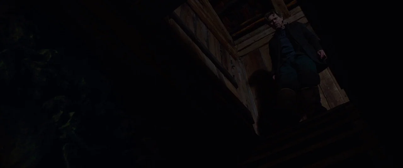 Siberia (2019) movie still: fullbody — A dimly lit narrow wooden staircase descends into heavy darkness. A man walks down the steep wooden steps…