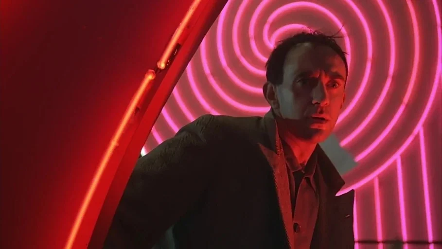 Brazil (1985) movie still: medium — A man stands before a wall featuring an illuminated pink neon spiral pattern. The man glances nervously to his…
