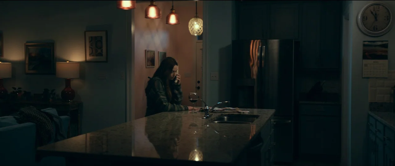 The Life of Chuck (2024) movie still: medium — A dark residential kitchen features a marble island with a glass of red wine. A young woman stands at…