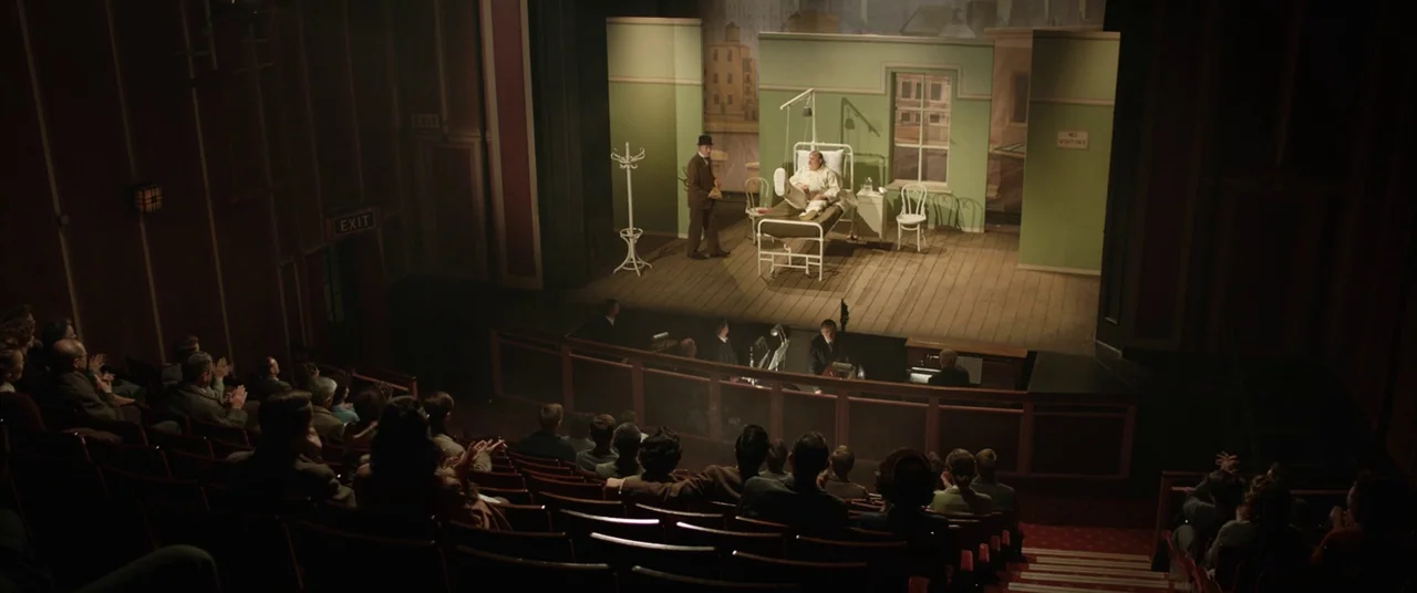 Domino (2005) movie still: wide — A theatrical stage displays a set designed as a hospital room with green walls. A man in a suit stands next to a…