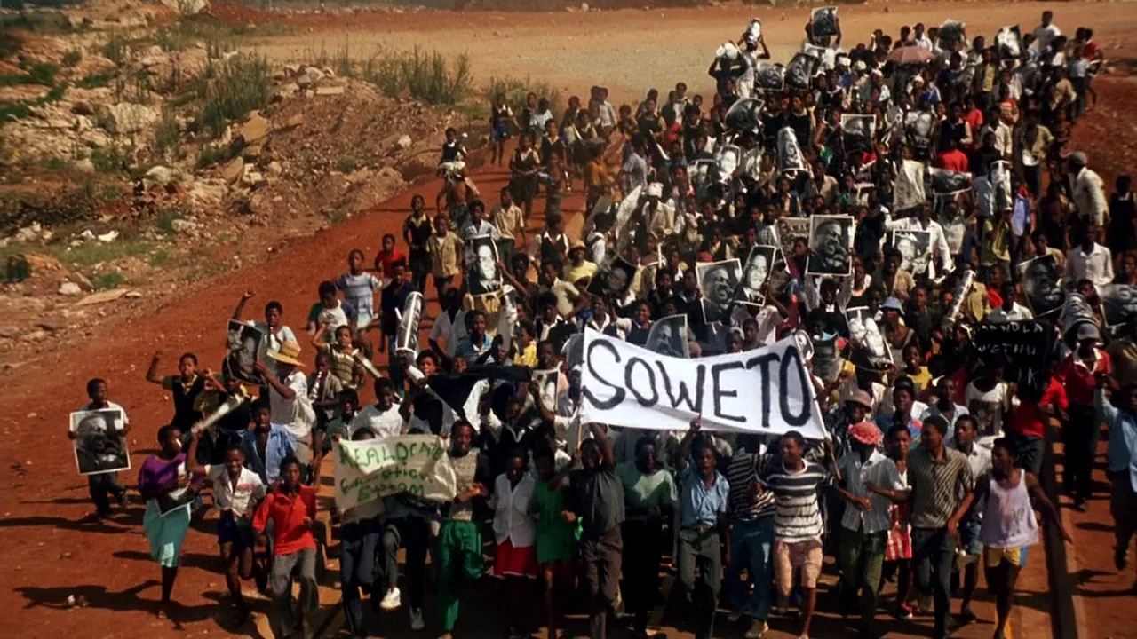 Malcolm X (1992) movie still: wide — A dense crowd of people marches down a desolate red dirt road. The protestors carry signs and banners with…