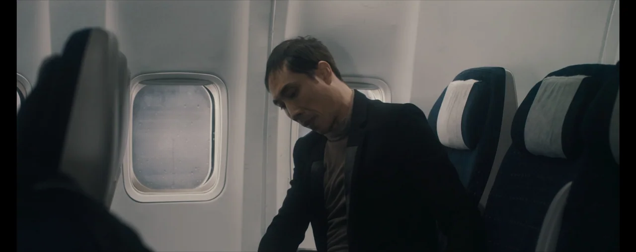 The Wrong Tenant (2022) movie still: medium — A man sits in an airplane seat beside a window with the shade up. He wears a black blazer and stares…