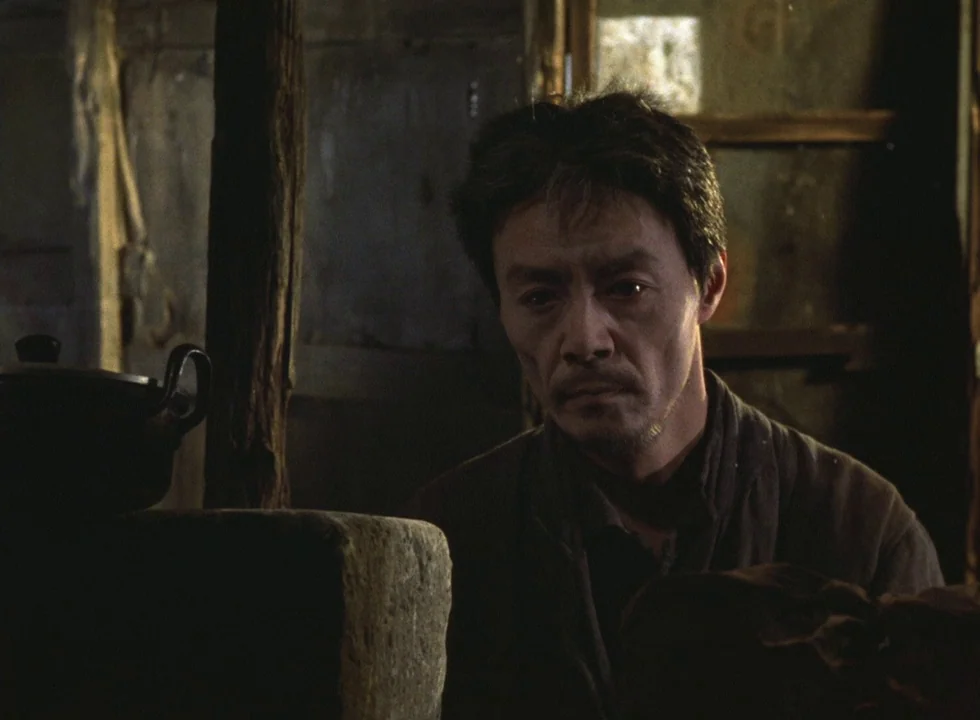 Dodes’ka-den (1970) movie still: medium — A dimly lit room features a wooden post and a dark ceramic pot. A man sits quietly looking forward in the…