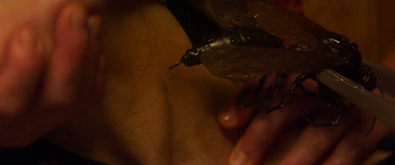 Mandy (2018) movie still: closeup — A dark indoor space focuses on a person's chest and shoulder area. A person uses metal tweezers to position a…
