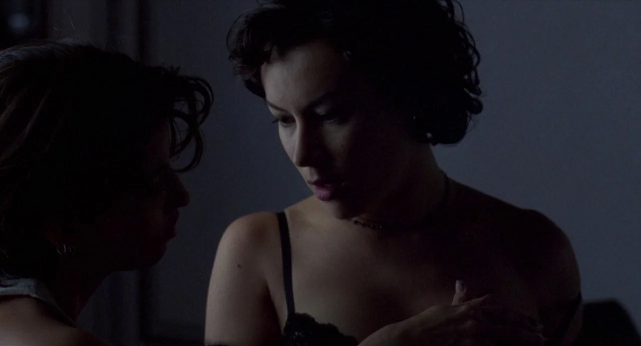 Bound (1996) movie still: medium — Two women stand in a dark room with minimal light reflecting on their skin. They are positioned facing each other…