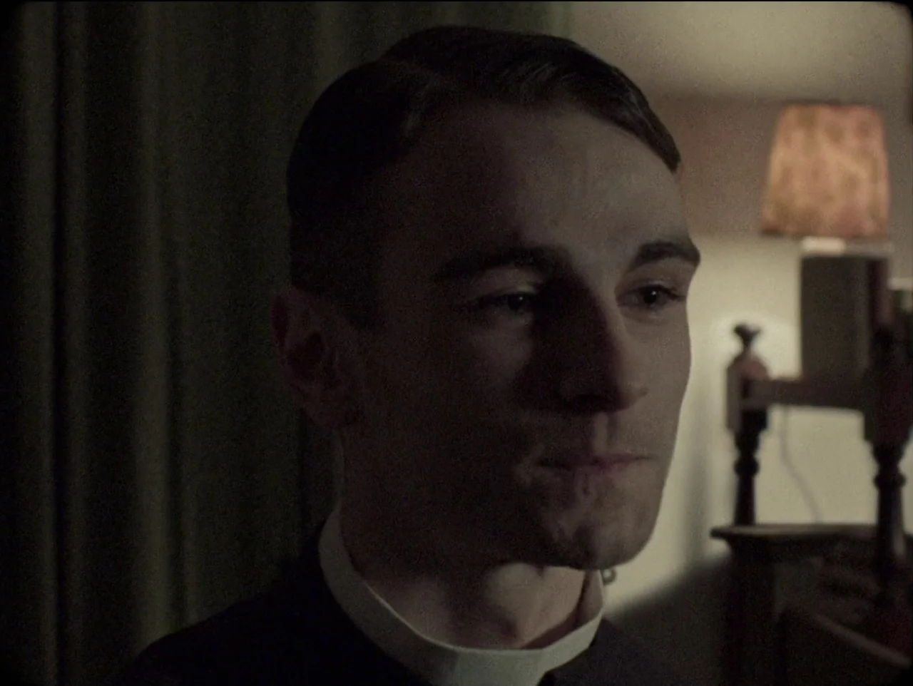 The Devil’s Doorway (2018) movie still: closeup — A dimly lit room contains a man wearing a black shirt with a clerical collar. The man remains…