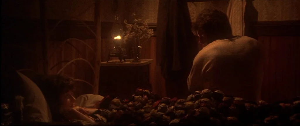 McCabe & Mrs.Miller (1971) movie still: medium — A dimly lit room with wooden wall paneling and a dark, heavy quilt. A man sits at the edge of the…